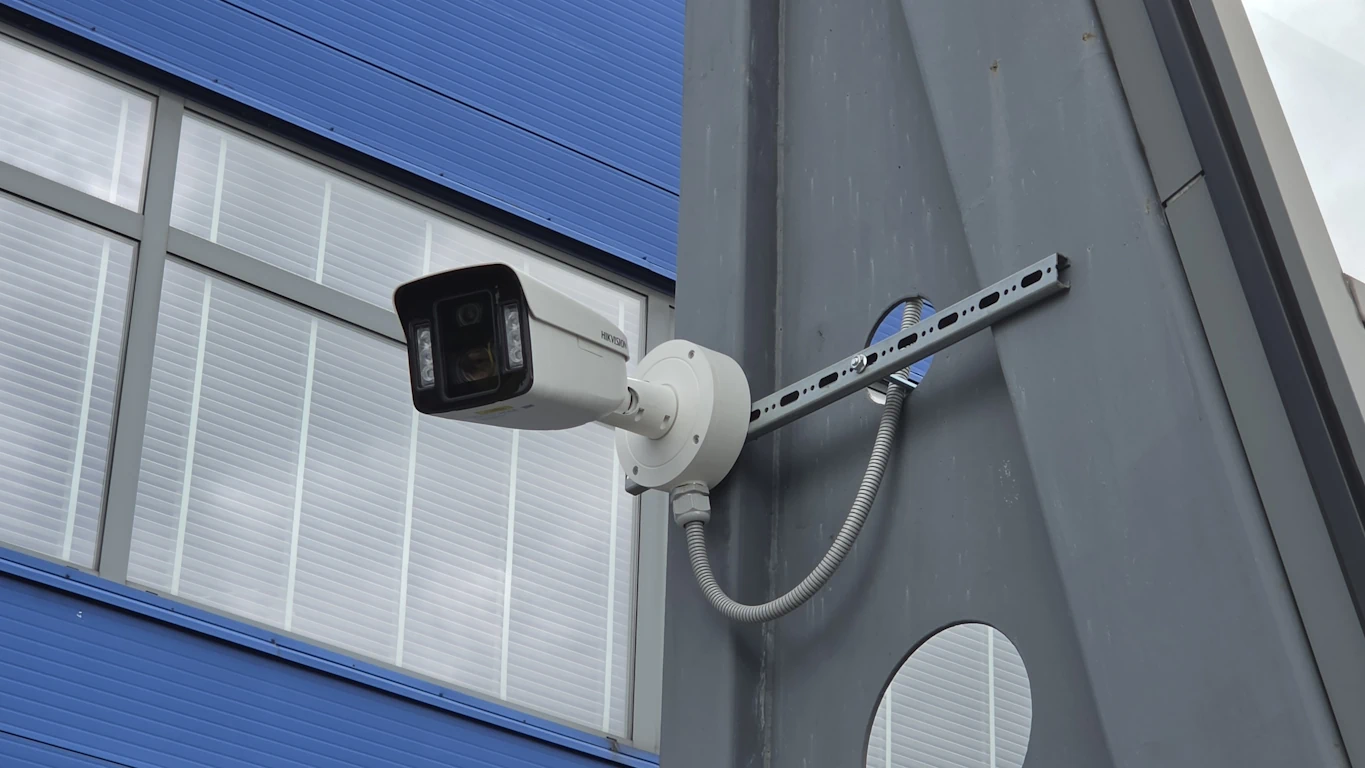 Zewnętrzna kamera monitoringu wizyjnego Hikvision zamontowana na szarym elemencie konstrukcyjnym na tle niebieskiej ściany IKEA